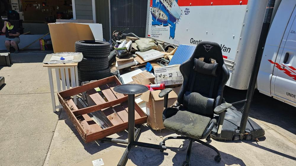 A driveway piled with an old desk chair, four tires, a broken planter box and flat-pack furniture beside a moving van