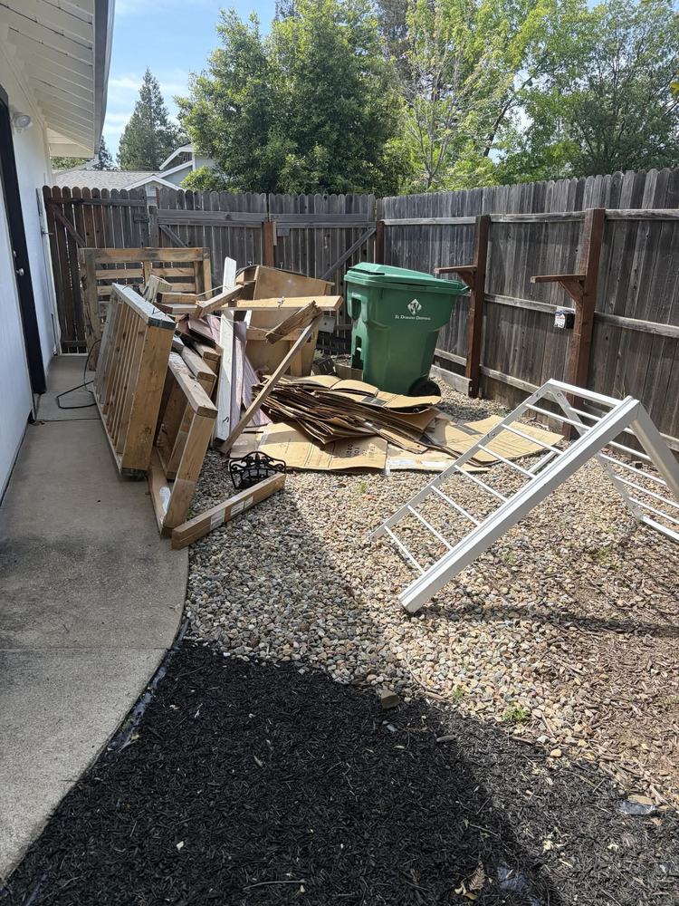 A side yard stacked with broken pallets, scrap lumber, flattened cardboard and an old screen door